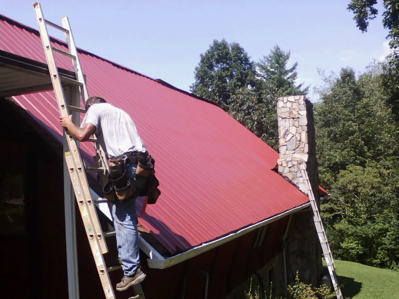 Roofer on a ladder beside a red metal roof and stone chimney — Soffit & Fascia Repair by Roofing Brothers in Greenwood
